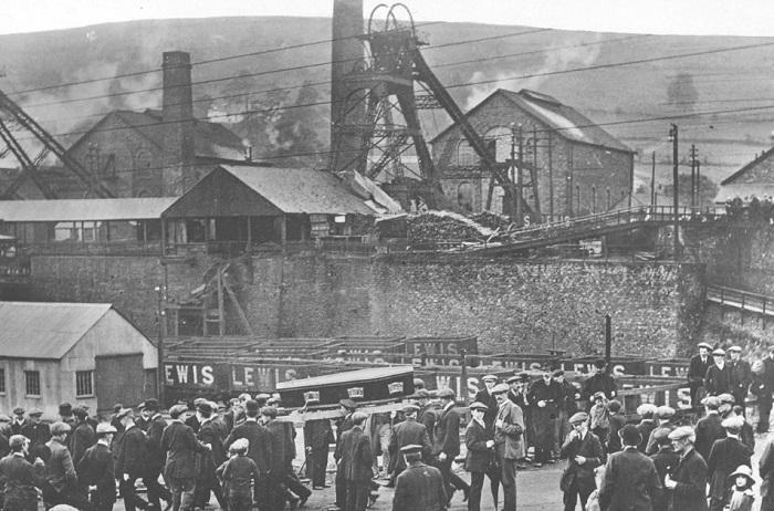 Black and white photograph, view of colliery with coffin being carried away.